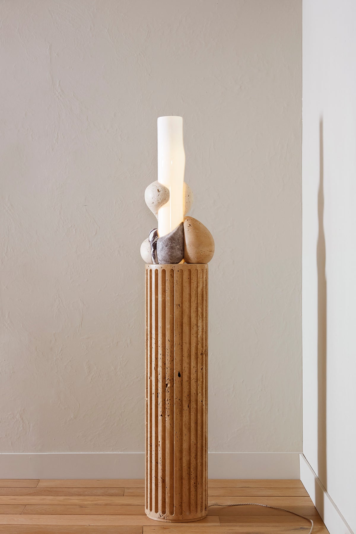 Decorative lamp with stone base and cylindrical glass structure surrounded by marble elements against a plain wall.