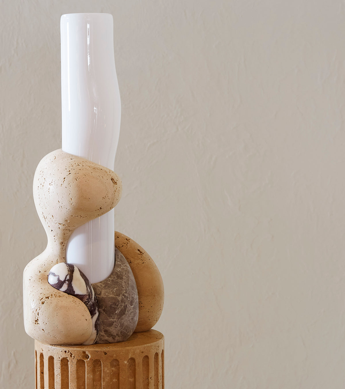 Decorative lamp with stone base and cylindrical glass structure surrounded by marble elements against a plain wall.