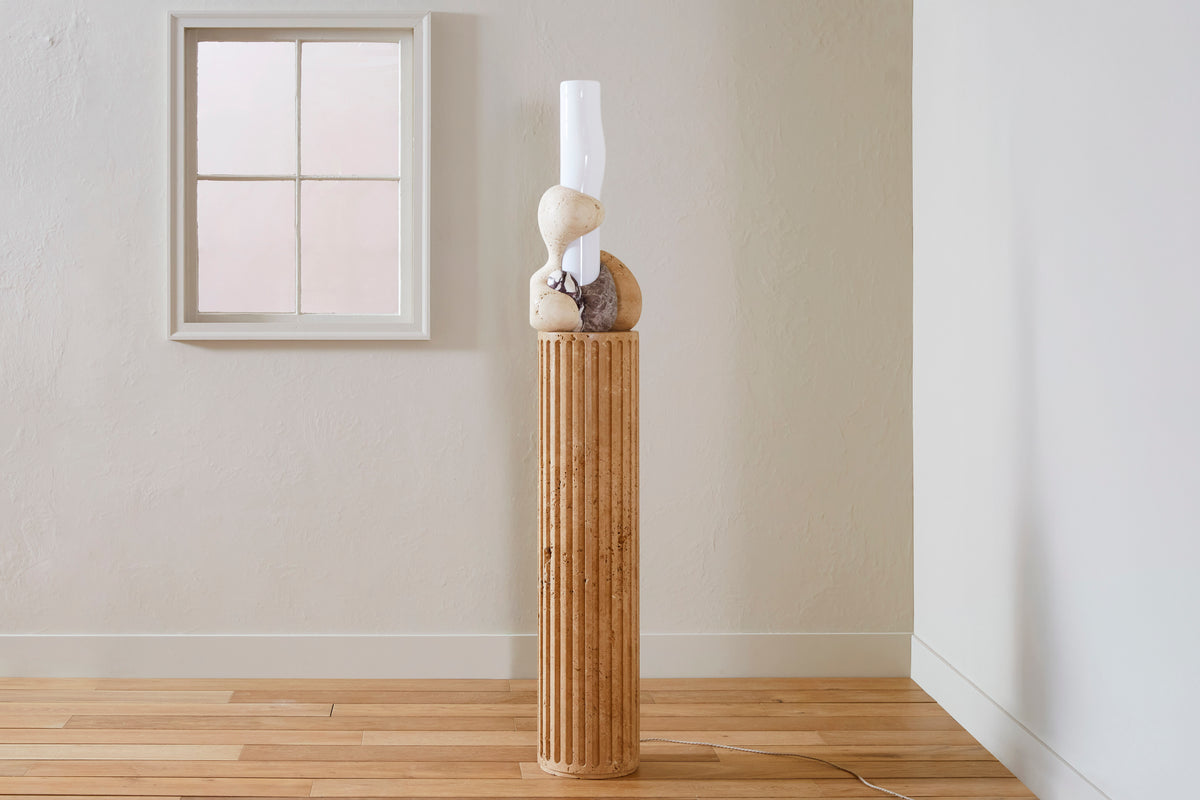 Decorative lamp with stone base and cylindrical glass structure surrounded by marble elements against a plain wall.